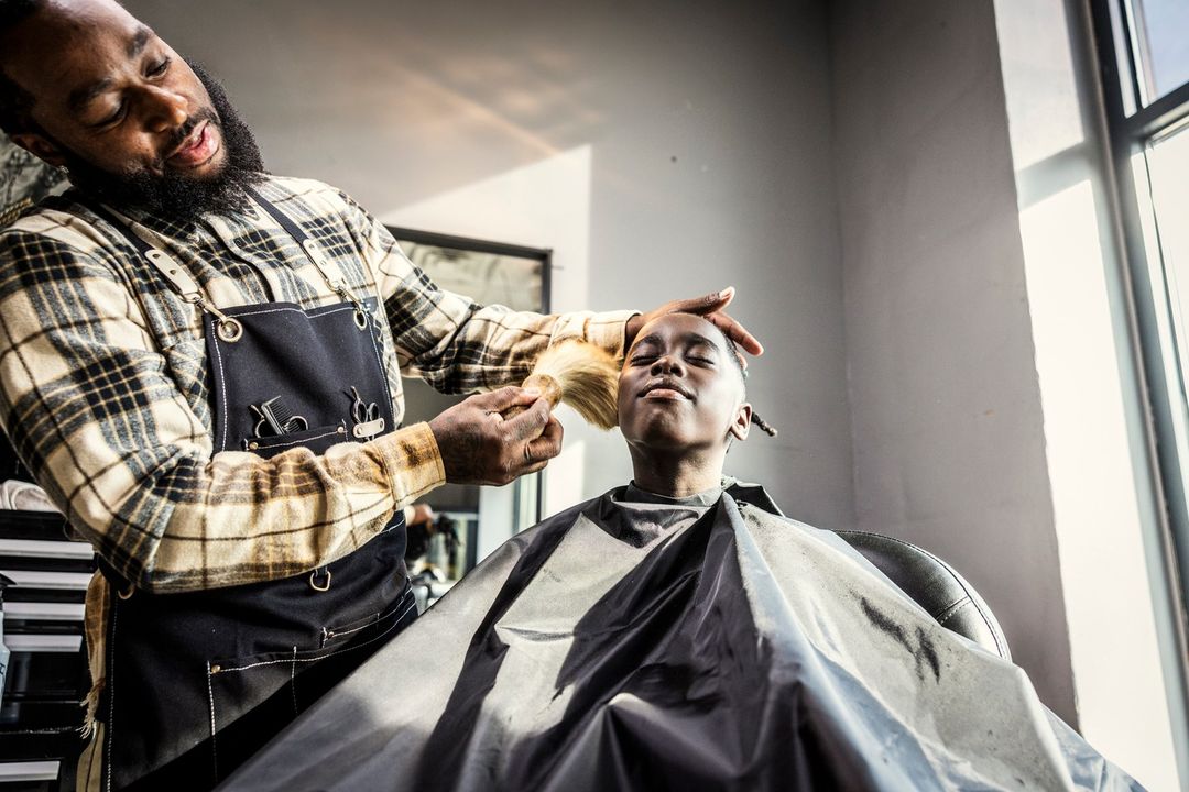 An African-American barber styles a young boy's hair in a bright barbershop, the barber wearing a plaid shirt and black apron.