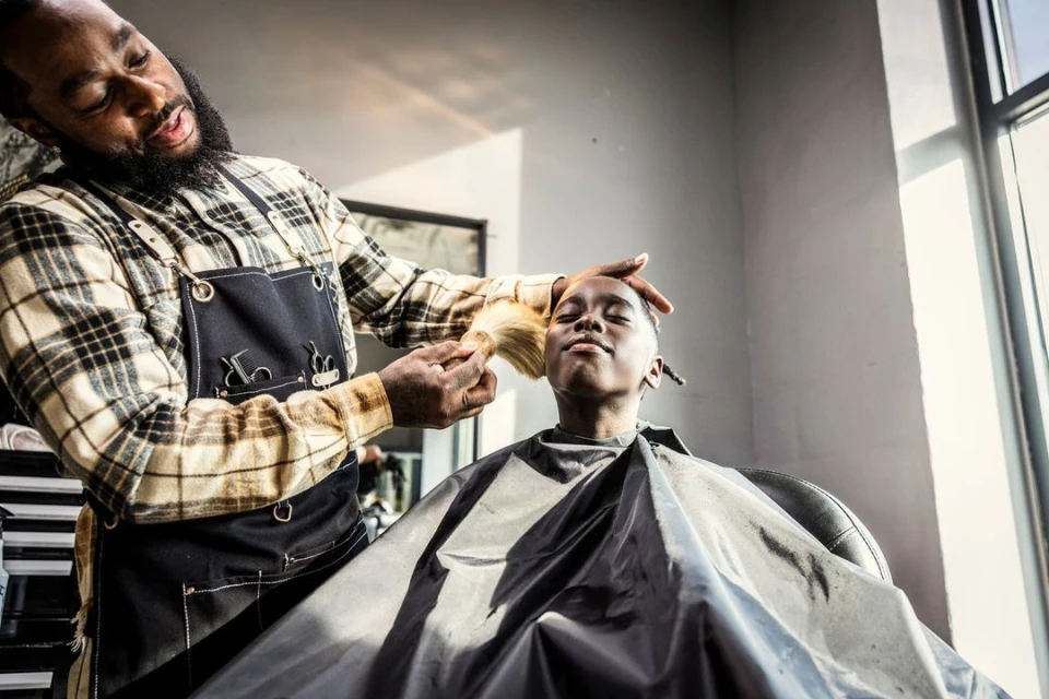 An African-American barber styles a young boy's hair in a bright barbershop, the barber wearing a plaid shirt and black apron.