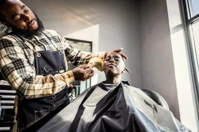 An African-American barber styles a young boy's hair in a bright barbershop, the barber wearing a plaid shirt and black apron.