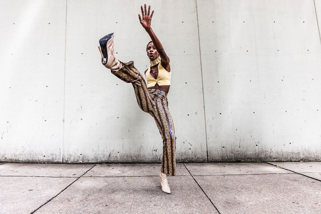 An adult female fashion model in patterned pants and high heels poses dynamically against a concrete wall in an outdoor setting.