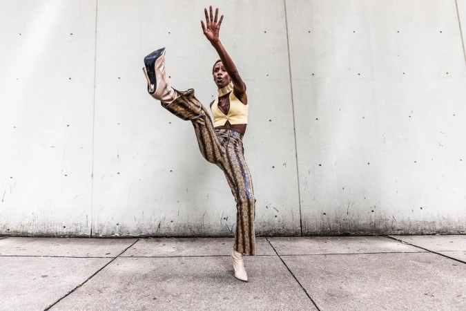 An adult female fashion model in patterned pants and high heels poses dynamically against a concrete wall in an outdoor setting.