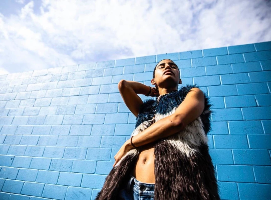 Portrait of a slim adult female posing outdoors against a turquoise brick wall with a blue and white sky. She's wearing a fur vest.