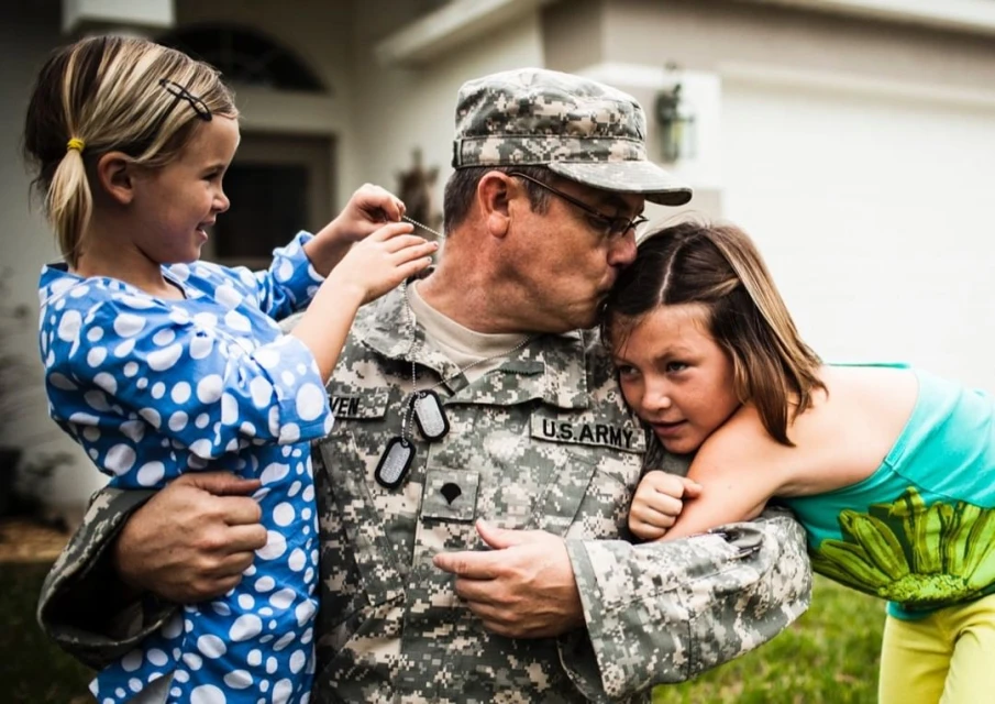 Military father hugs his two daughters, being kissed on the forehead in his camouflage uniform outside their home on a sunny day.