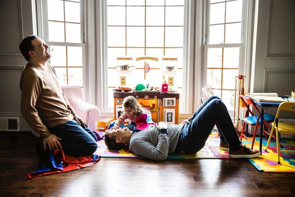 Father and daughter play together on a colorful mat in a brightly lit living room next to a play kitchen and other toys.