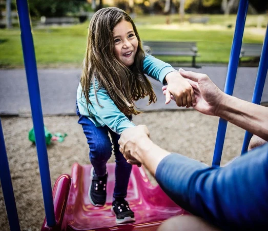 A young girl climbs a playground slide with help from an adult, set in a park with green grass and trees in the background.