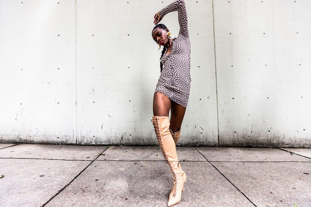 Model in patterned minidress and tall beige laced boots posing against concrete wall, photographed by Pat Molnar