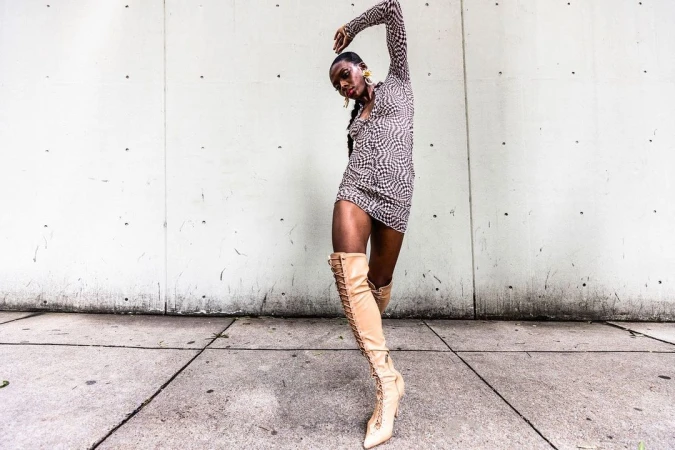 Model in patterned minidress and tall beige laced boots posing against concrete wall, photographed by Pat Molnar