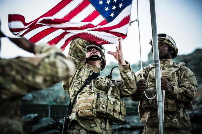 Three soldiers in camouflage uniforms raise the American flag on a metal pole outdoors in a rugged, rocky environment under cloudy skies.