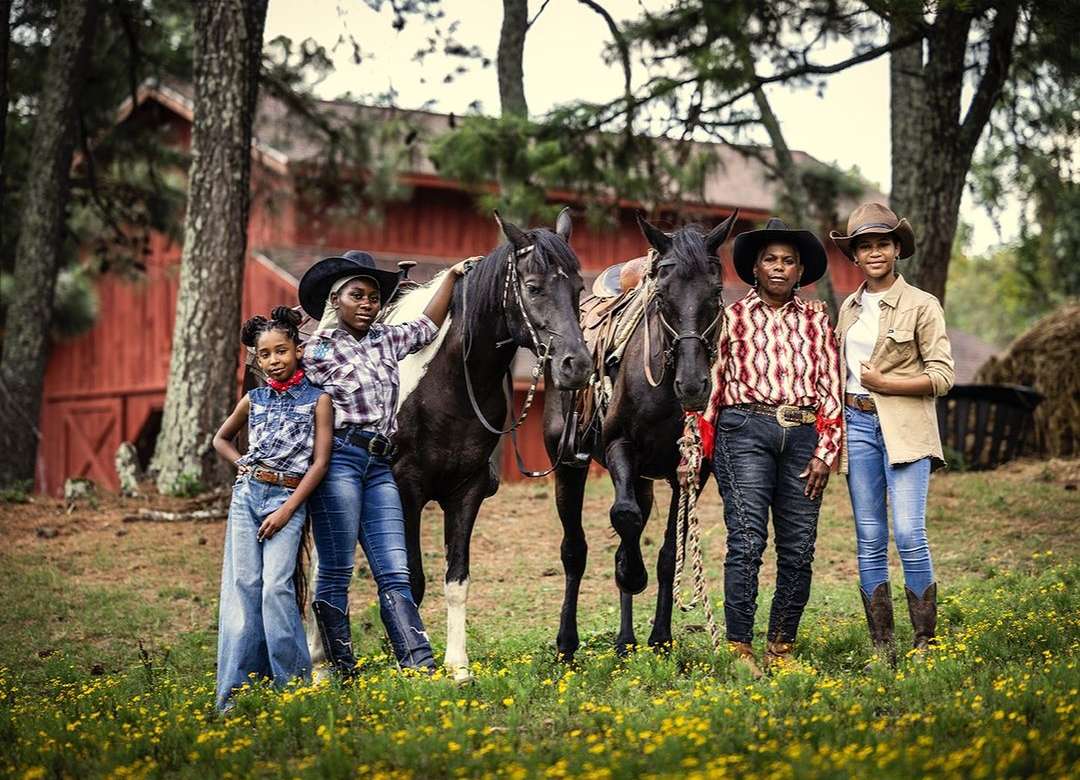 Group of four people, two horses standing in a field of yellow flowers, wearing cowboy hats, red barn and trees in background.