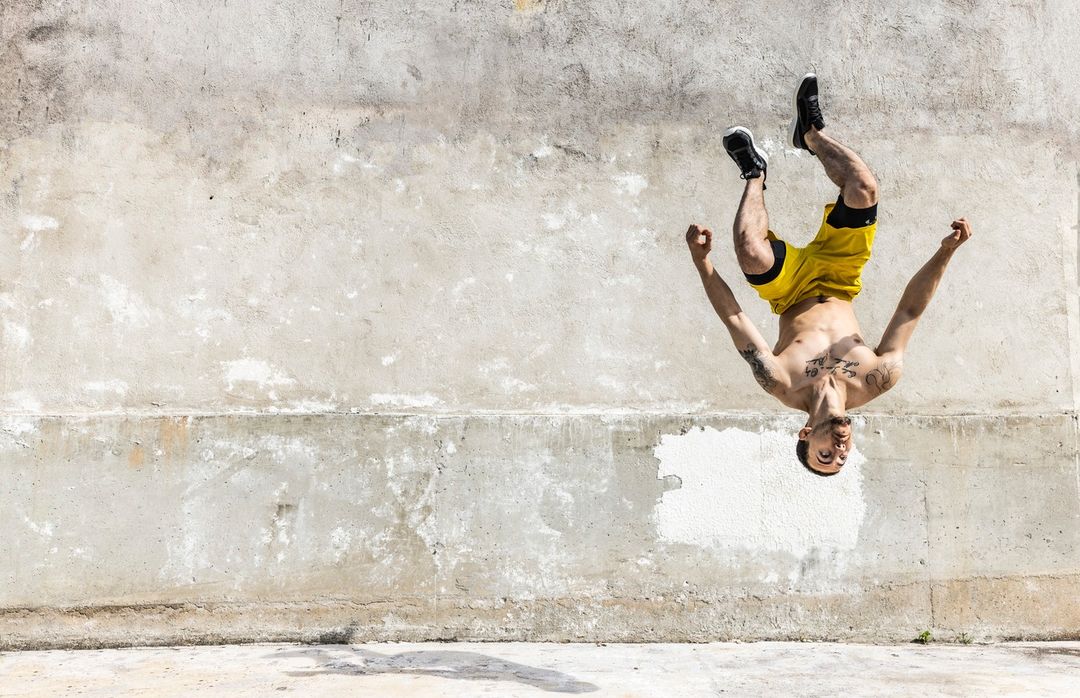 Athletic man performing a backflip against a textured concrete wall outdoors, wearing yellow shorts and sneakers, captured in daylight.