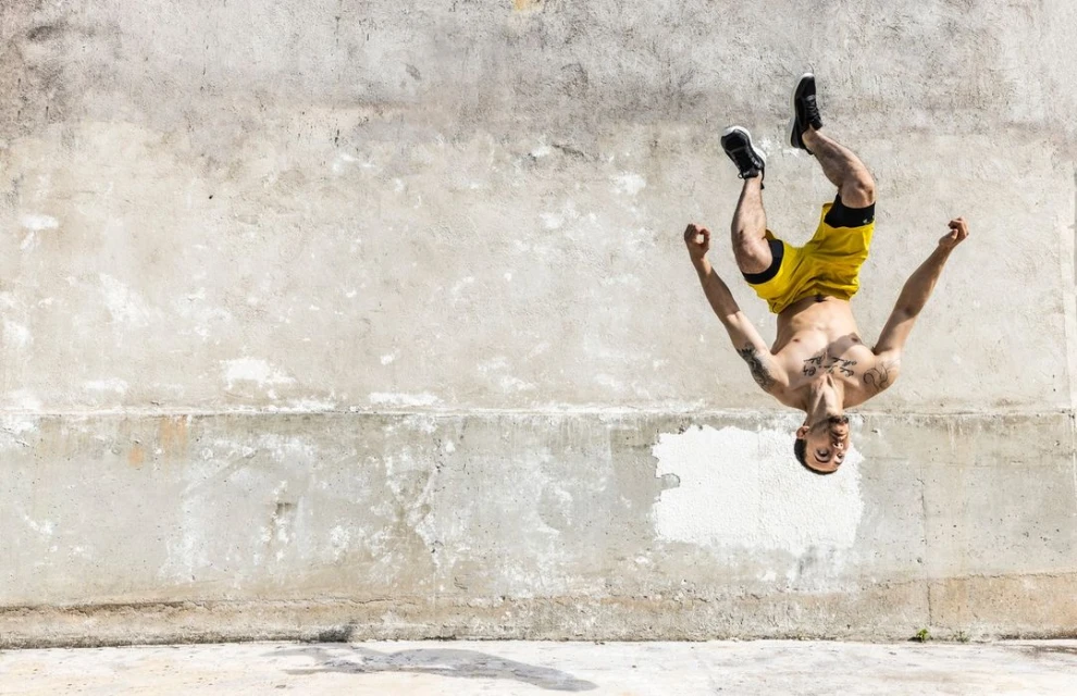 Athletic man performing a backflip against a textured concrete wall outdoors, wearing yellow shorts and sneakers, captured in daylight.