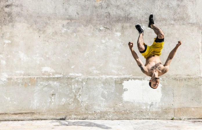 Athletic man performing a backflip against a textured concrete wall outdoors, wearing yellow shorts and sneakers, captured in daylight.