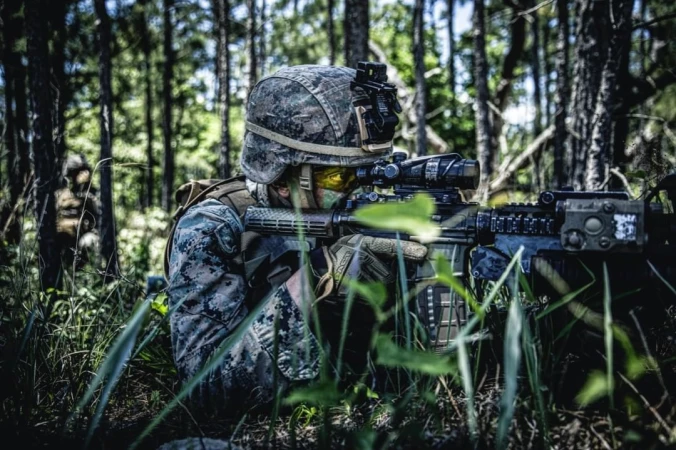 Camouflaged soldier in military gear kneels in a forest setting holding an assault rifle with optical sight, surrounded by green foliage.