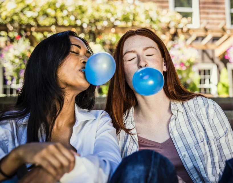 Two young women outdoors blowing large blue bubblegum bubbles, wearing casual clothing against a natural, sunny, green background.