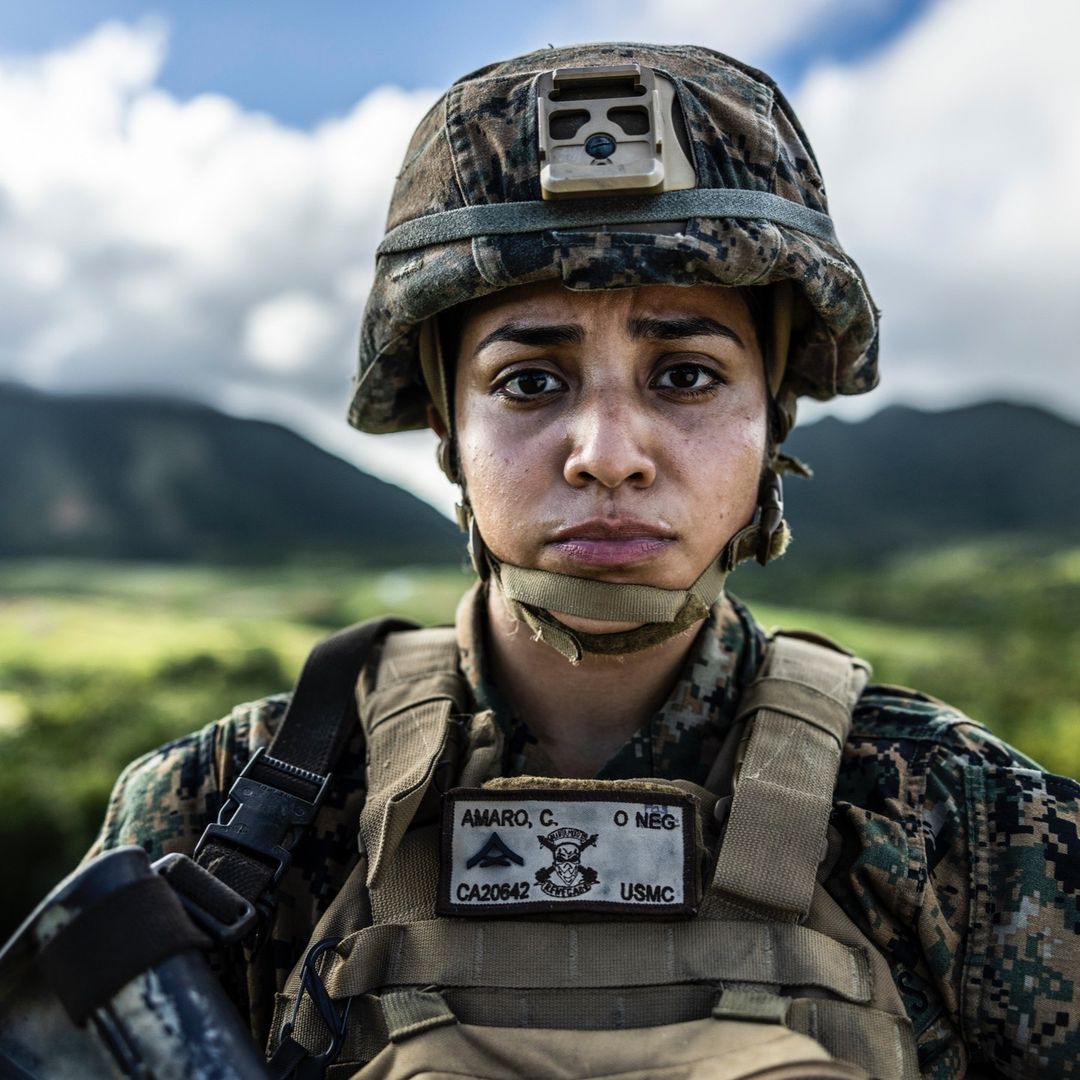 Close-up portrait of a young adult female in a military uniform and helmet outdoors with a mountain range in the background on a sunny day.