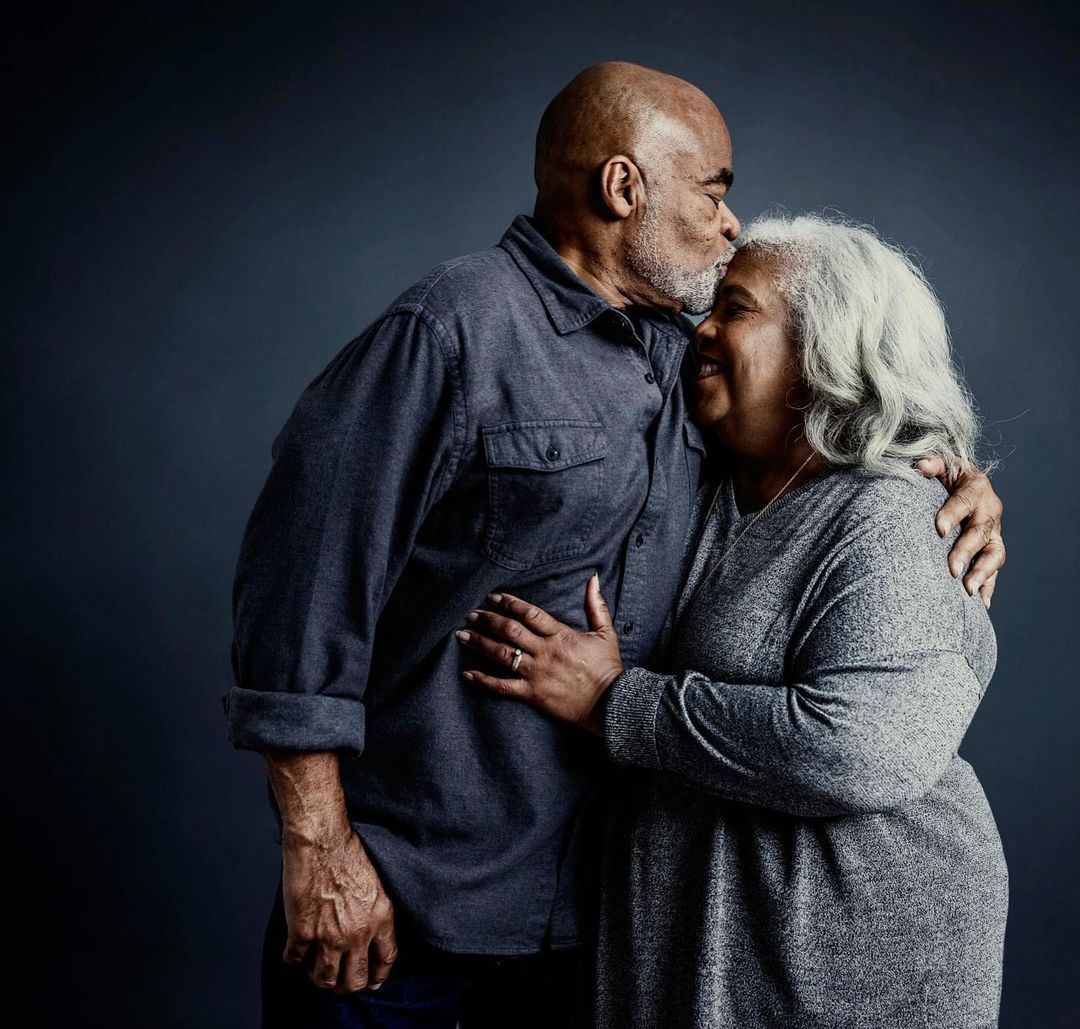 Affectionate moment of an African-American elderly couple, with the man kissing the woman's forehead in a loving embrace against a gray backdrop.