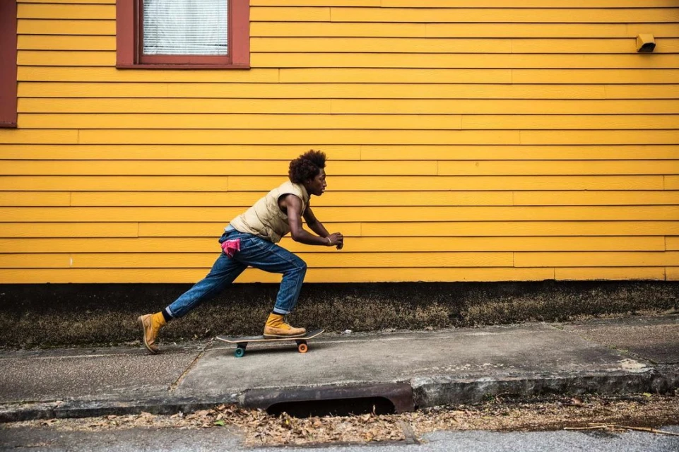 Person skateboarding on a sidewalk next to a yellow building. The skateboarder is wearing a vest and jeans, captured in motion.