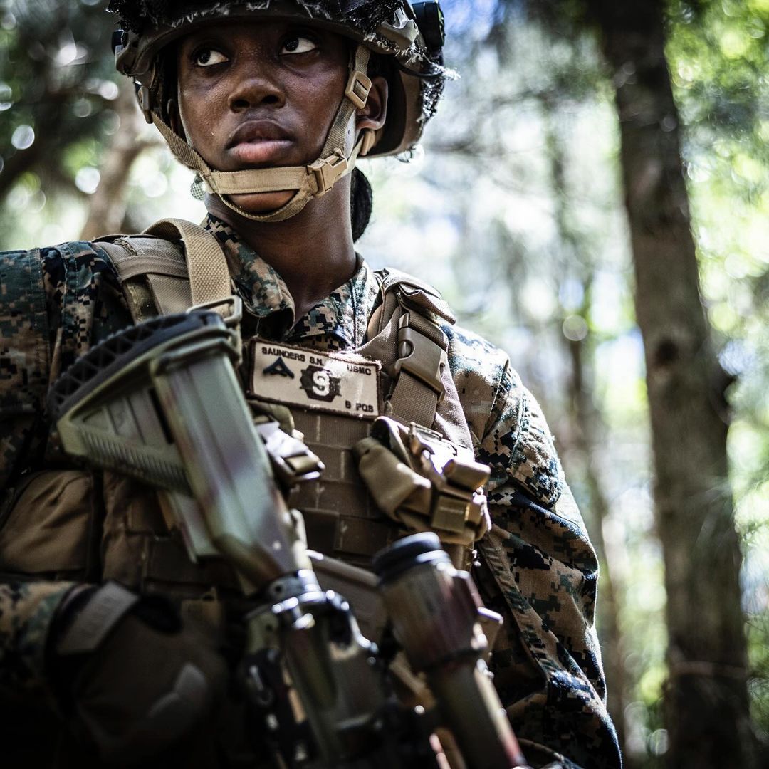 Woman in camouflage uniform with rifle in outdoor forest setting. Soldier or military personnel in action, medium shot.