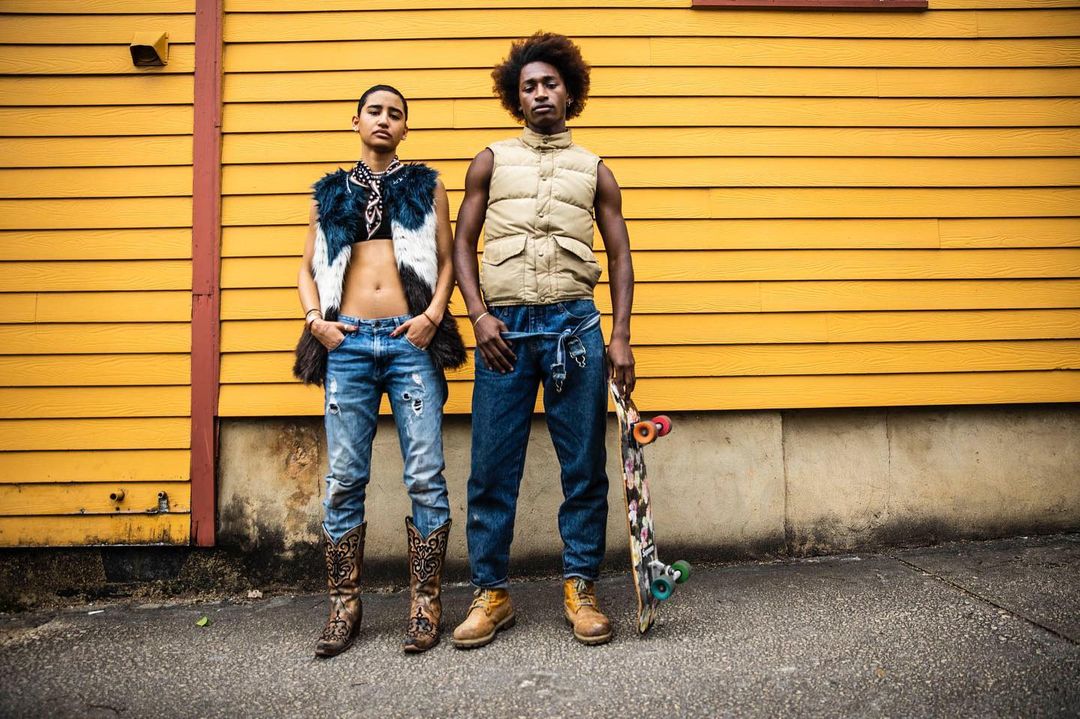 Two young adults strike a pose outdoors with a skateboard against a yellow-painted building in stylish street fashion.