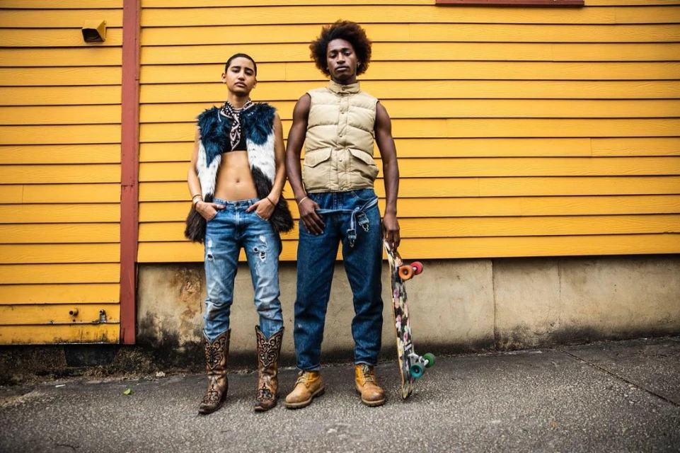 Two young adults strike a pose outdoors with a skateboard against a yellow-painted building in stylish street fashion.