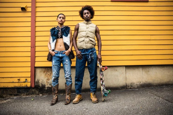 Two young adults strike a pose outdoors with a skateboard against a yellow-painted building in stylish street fashion.