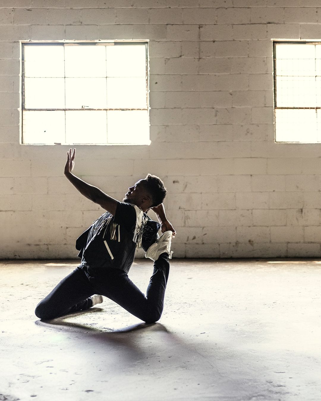 An athletic black woman performs a yoga pose indoors, illuminated by bright windows. The shot captures movement, flexibility, and natural light.