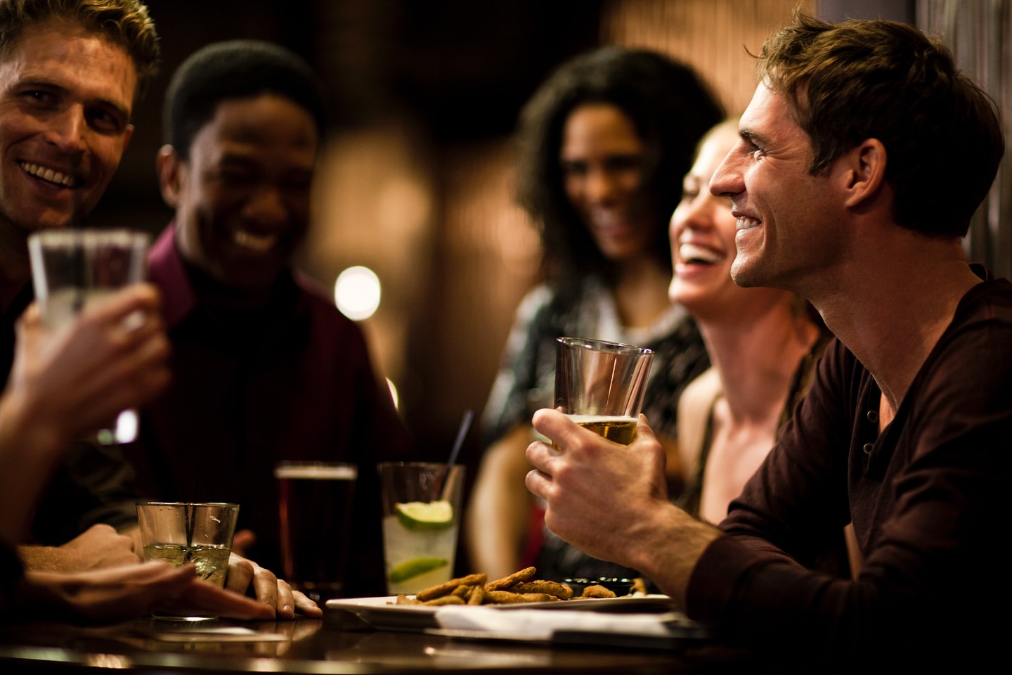 Group of friends laughing and socializing at a bar with drinks and snacks on the table in a dimly lit, social atmosphere.