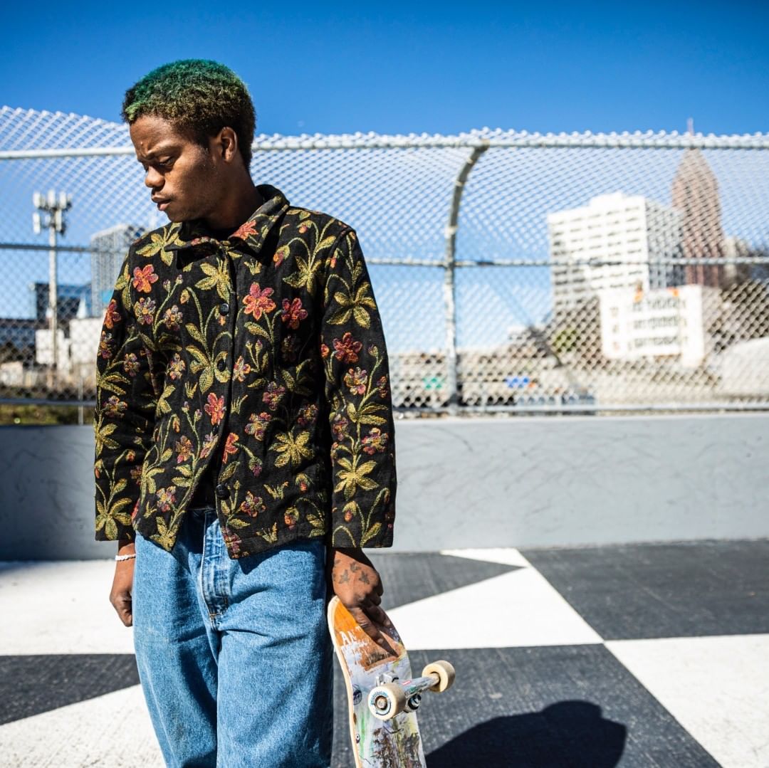 Young man with green hair in a patterned jacket holding a skateboard, standing in an outdoor urban setting with cityscape background.