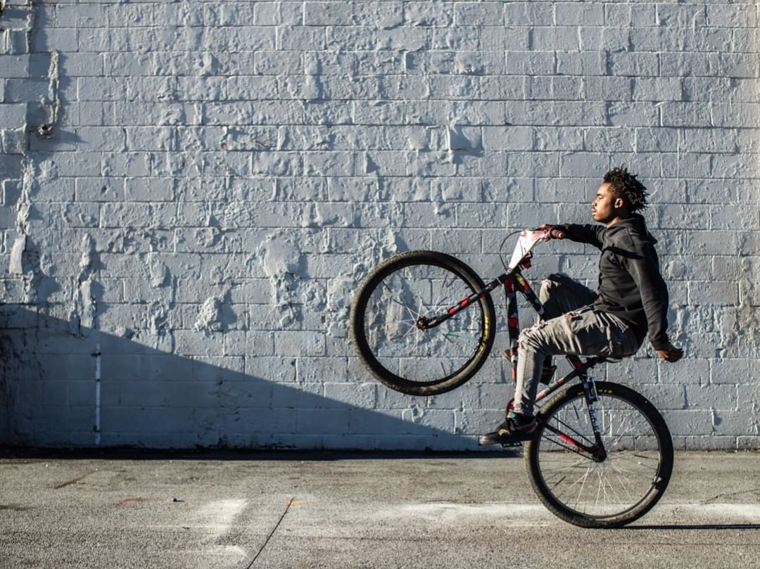 Adult male performing a wheelie on a bicycle, in front of a concrete brick wall on an asphalt surface on a sunny day.
