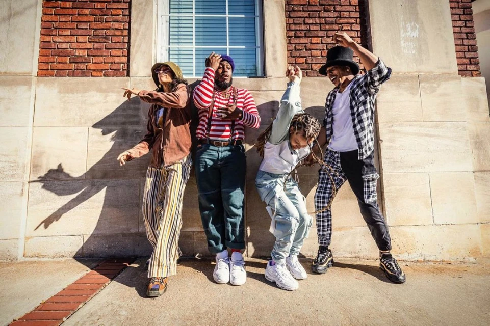 Four people pose against a brick wall with a window, showcasing diverse streetwear styles with a mix of colors, patterns, and textures.