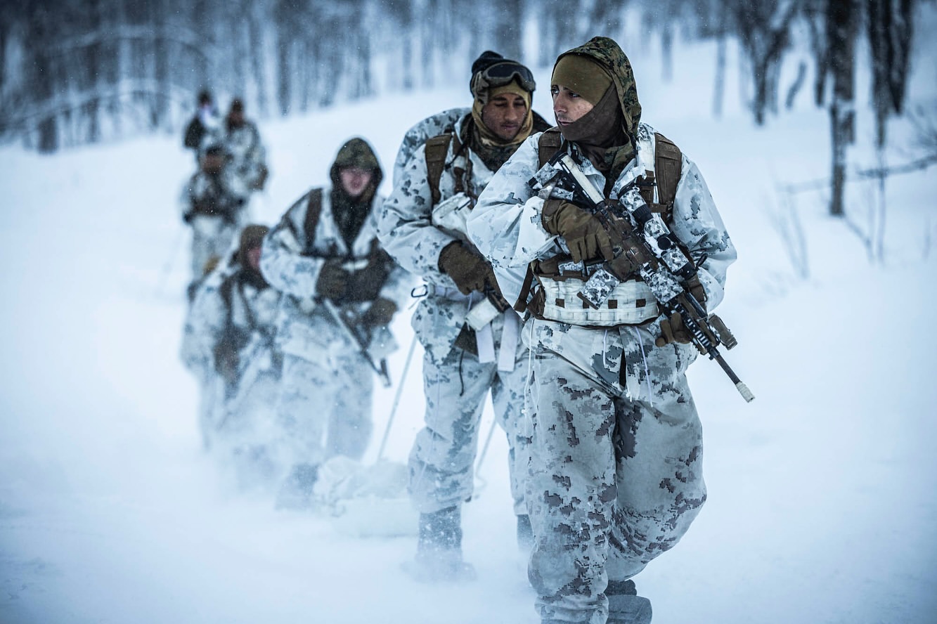 A group of armed soldiers in camouflage winter gear march through a snow-covered forest, carrying rifles and pulling sleds in daylight.