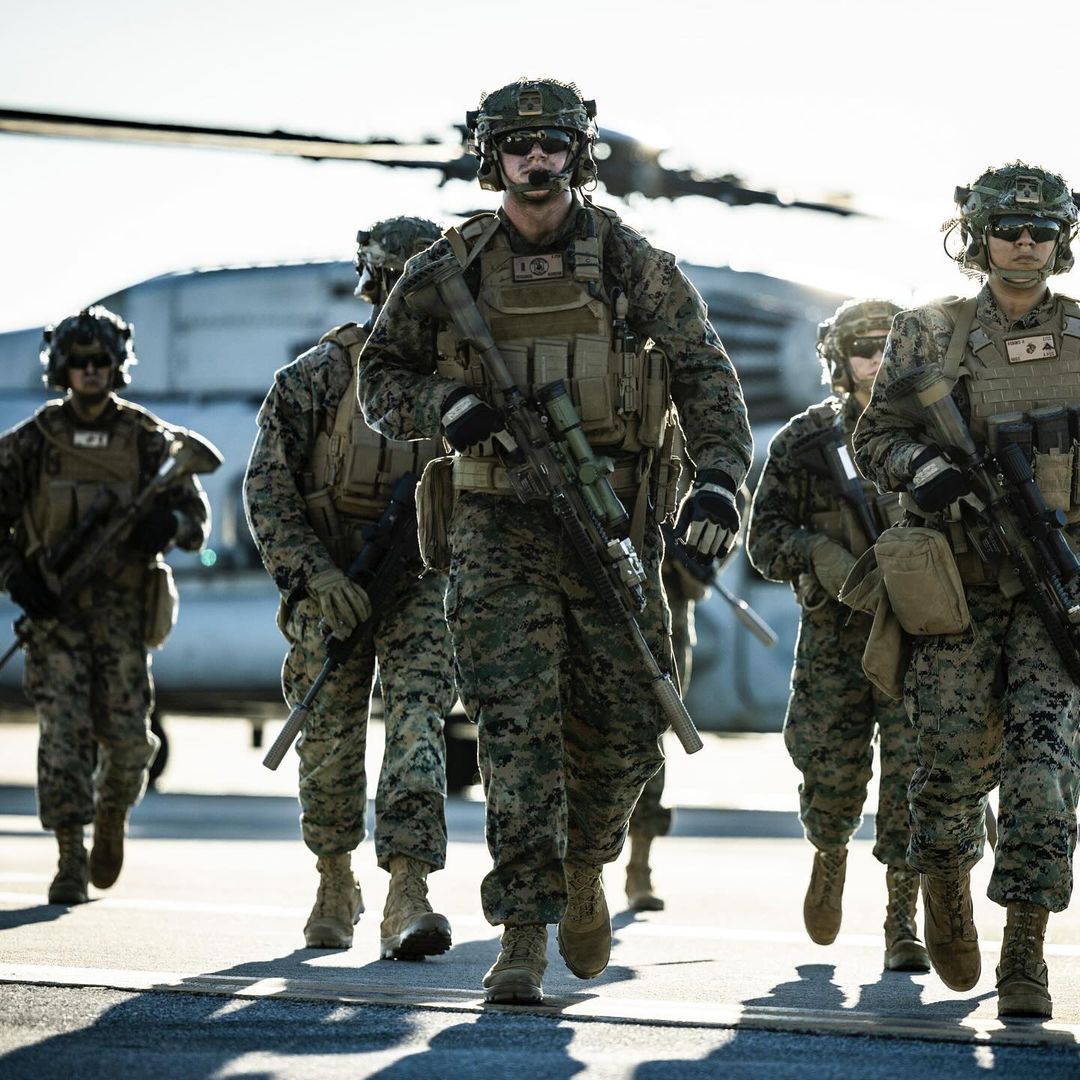 Military personnel in camouflage uniforms walk in formation near a helicopter on a tarmac, focusing on the teamwork and tactical gear.