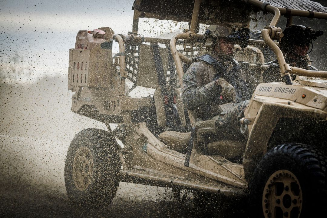 Two soldiers in military vehicle drive through mud. Tan tactical vehicle splashes in dirt; one adult male soldier drives through rough terrain.