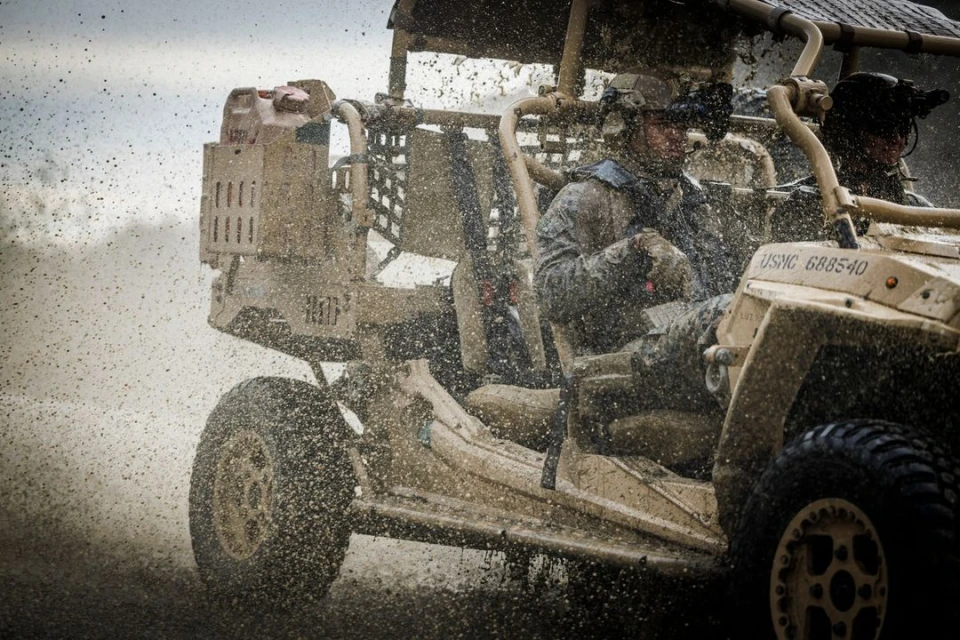 Two soldiers in military vehicle drive through mud. Tan tactical vehicle splashes in dirt; one adult male soldier drives through rough terrain.