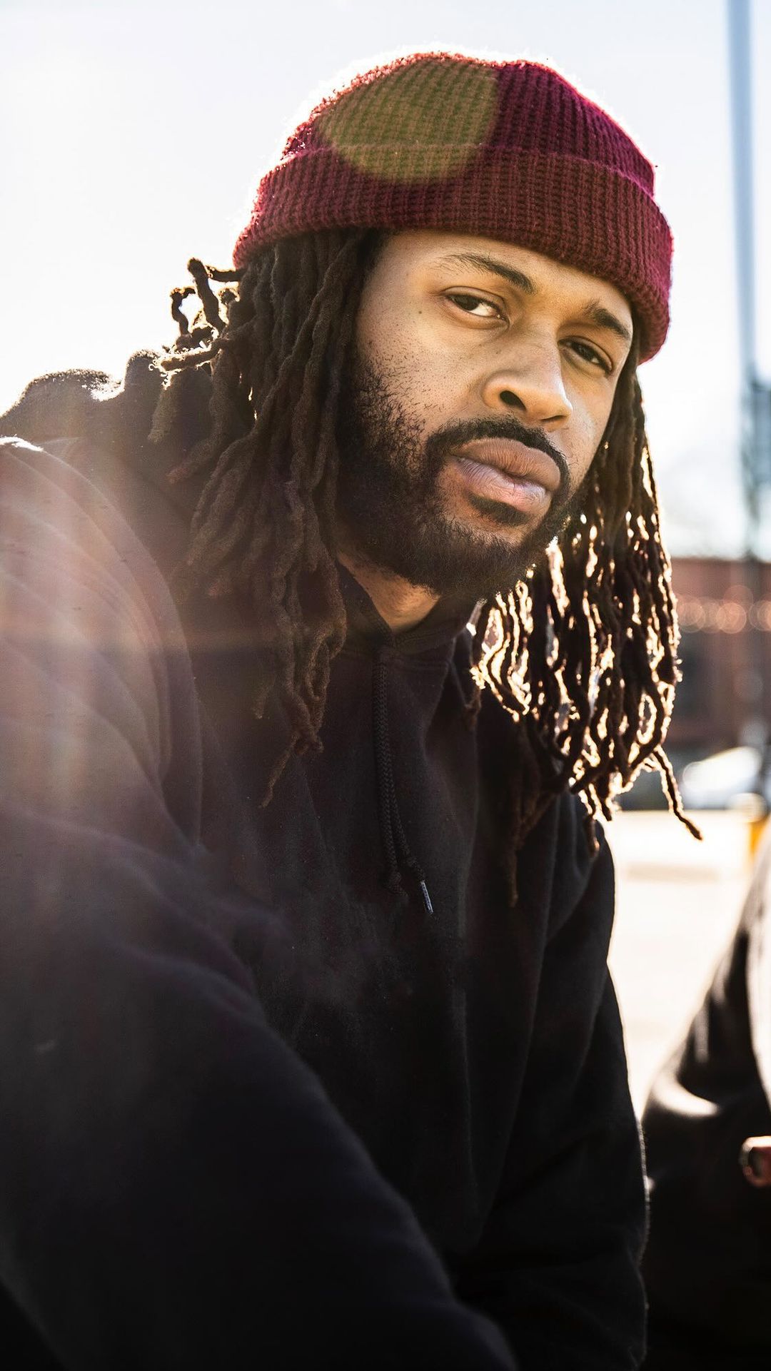 Portrait of a man with dreadlocks wearing a dark hoodie and maroon beanie in an outdoor setting with a blurred background.