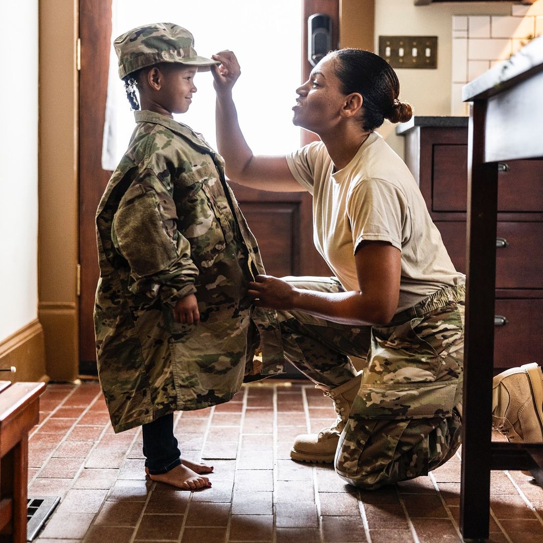 A child is wearing a camouflage military uniform with a woman helping adjust their hat in an indoor setting with warm lighting.