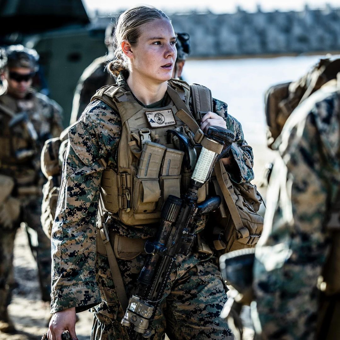 Female marine in camouflage uniform and tactical vest carrying rifle with blurred background of other military personnel and outdoor setting.