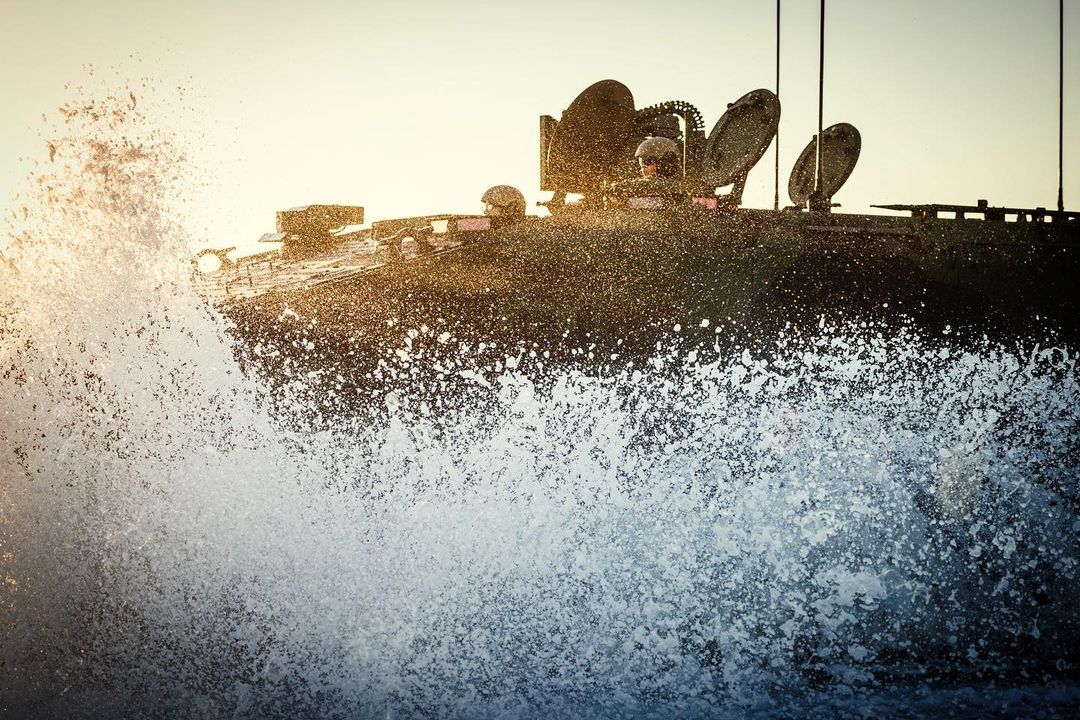 Military tank splashes through water with soldiers during daytime, featuring marine warfare and dramatic water effects.