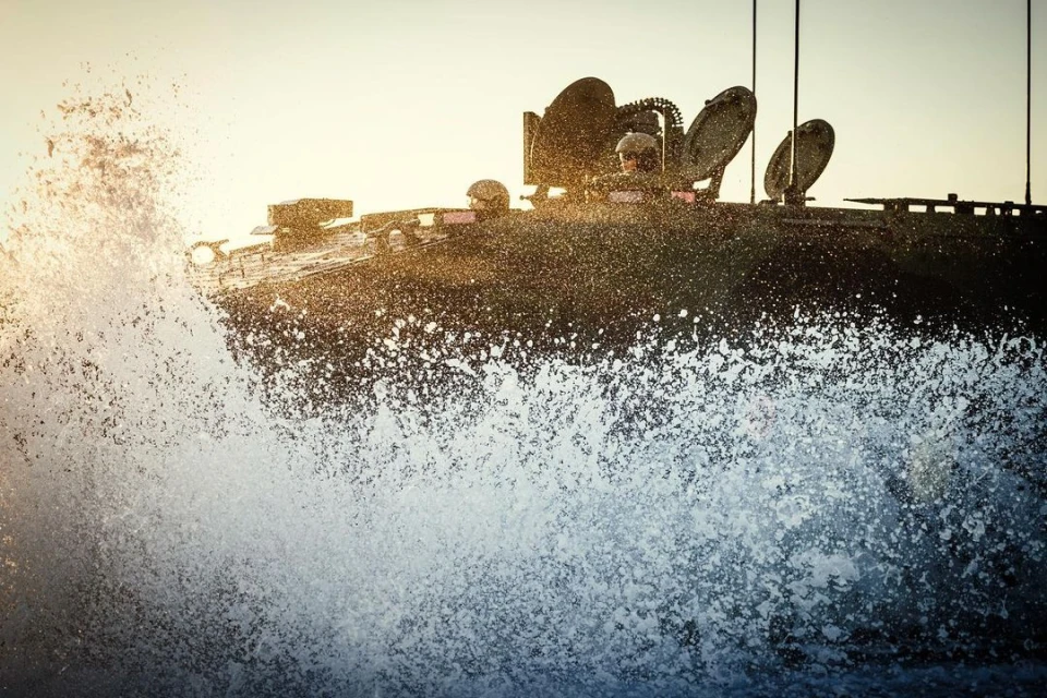 Military tank splashes through water with soldiers during daytime, featuring marine warfare and dramatic water effects.