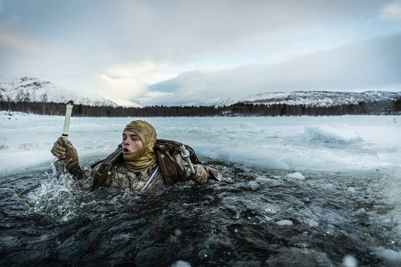 Adult in camouflage gear emerges from icy water, holding a white object, set against a backdrop of snowy mountains and cloudy sky.