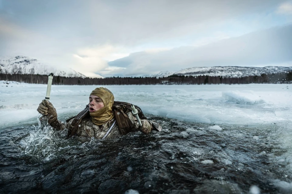 Adult in camouflage gear emerges from icy water, holding a white object, set against a backdrop of snowy mountains and cloudy sky.