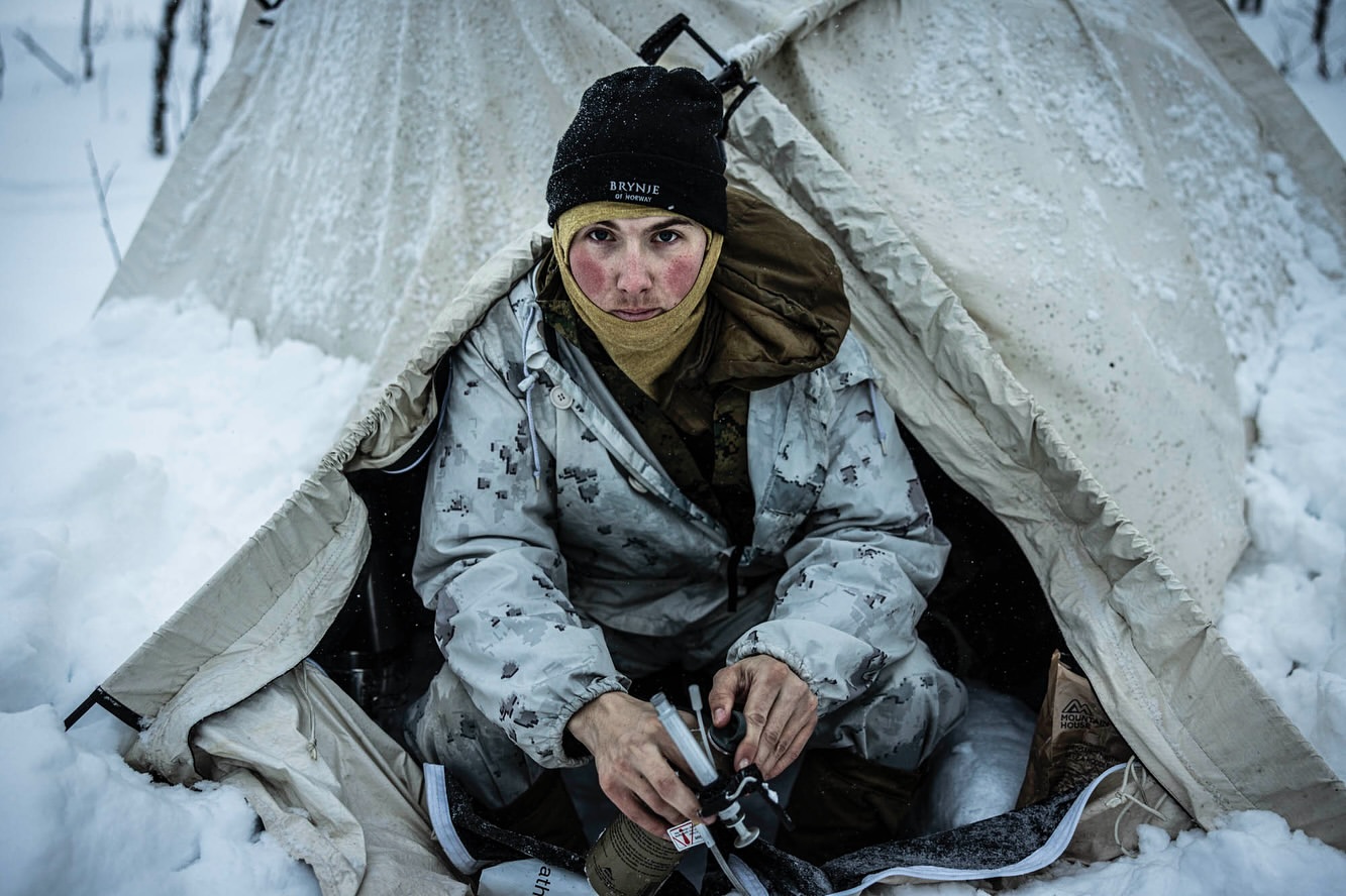 Outdoor portrait of a person in a white camouflage suit and black beanie inside a canvas tent surrounded by snow.