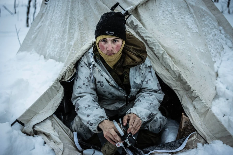 Outdoor portrait of a person in a white camouflage suit and black beanie inside a canvas tent surrounded by snow.