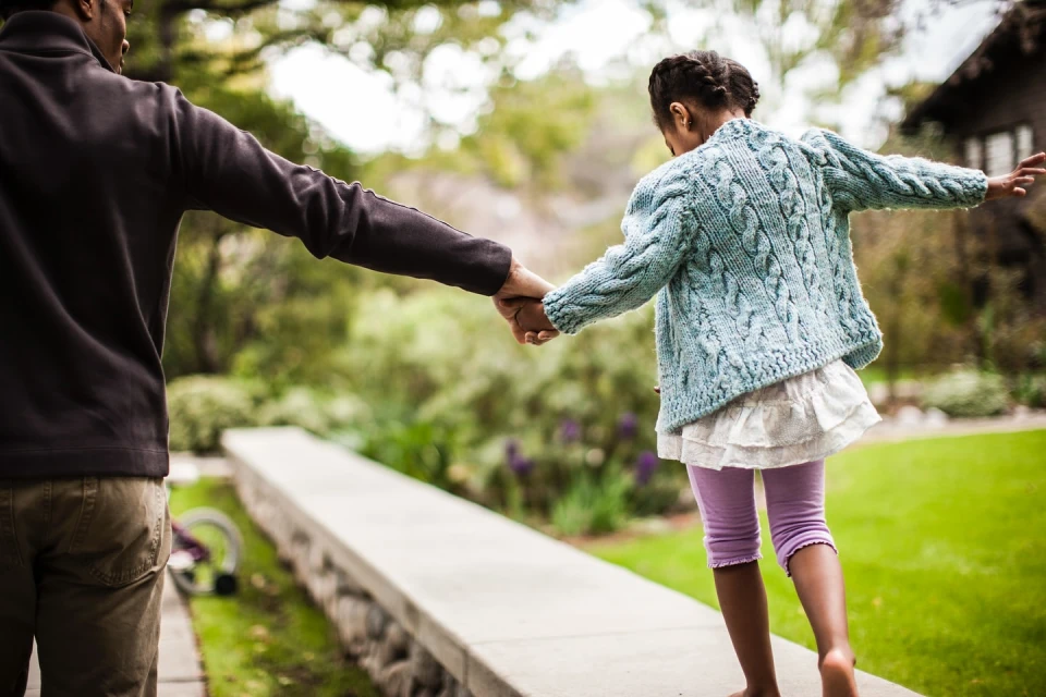 Father and daughter holding hands while balancing on a concrete ledge in a green, outdoor setting, showcasing family and quality time.
