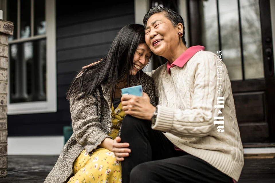 Asian mother and daughter embrace on porch steps. Smiling, faces close, wearing sweaters. House visible in background, natural light.