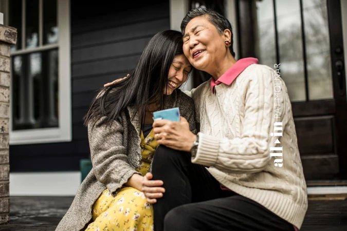 Asian mother and daughter embrace on porch steps. Smiling, faces close, wearing sweaters. House visible in background, natural light.
