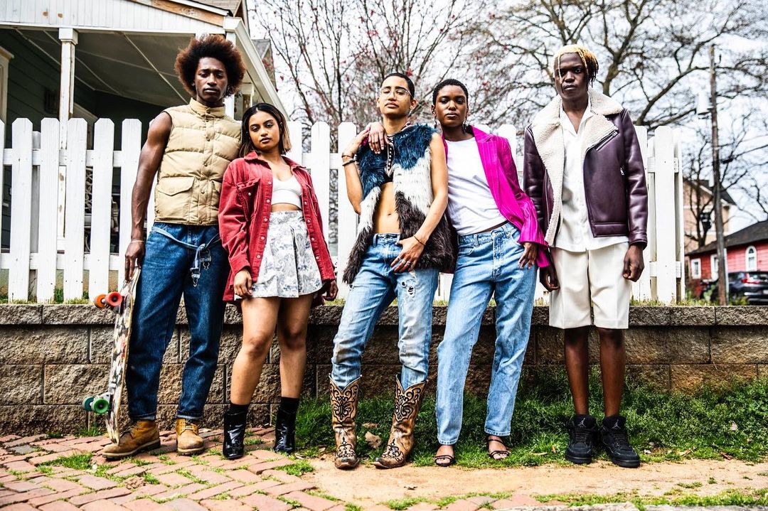 Group of five young adults stand together in an outdoor urban setting wearing stylish casual clothing; a house with a white fence visible.