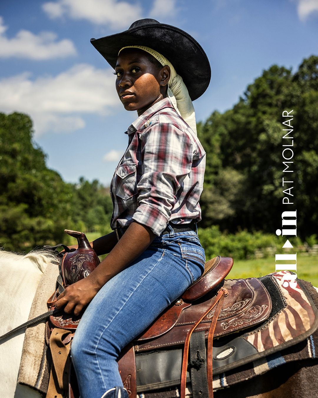 Portrait of a young Black woman in a cowboy hat and plaid shirt, riding a white horse on a sunny day, with green trees in the distance.