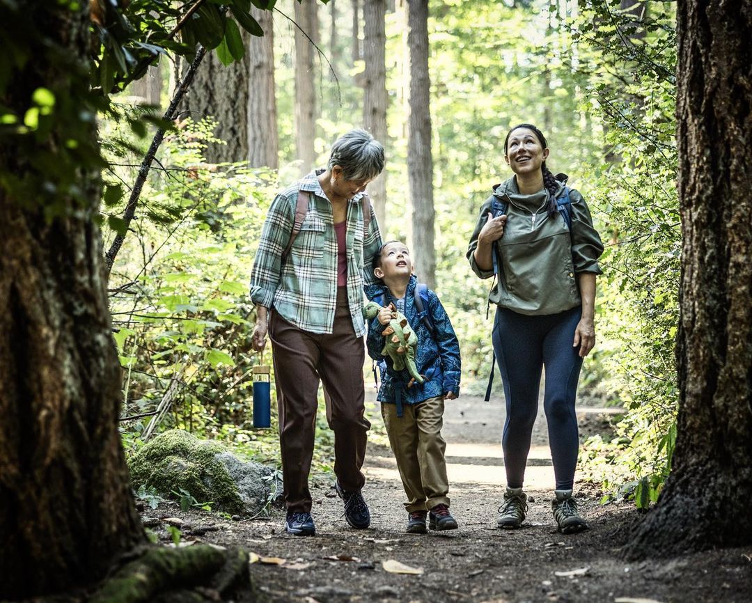 Three people walk along a dirt path through a sun-dappled forest: an older woman, a young child holding a toy dinosaur, and an adult woman.