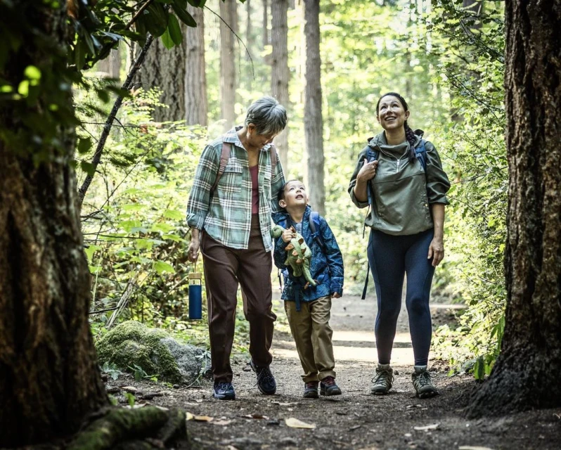 Three people walk along a dirt path through a sun-dappled forest: an older woman, a young child holding a toy dinosaur, and an adult woman.